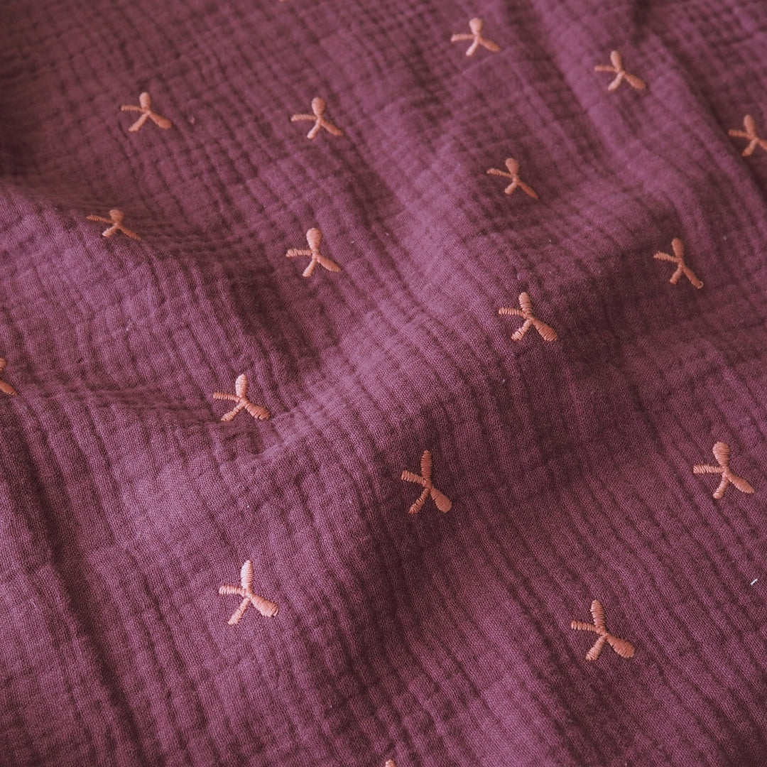 A close-up of a textured, crinkled gauze fabric in a dusty plum purple color, decorated with a scattered pattern of small, embroidered peach-pink bow motifs.