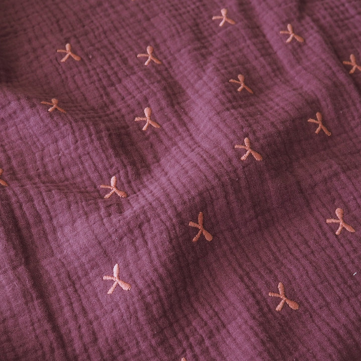 A close-up of a textured, crinkled gauze fabric in a dusty plum purple color, decorated with a scattered pattern of small, embroidered peach-pink bow motifs.
