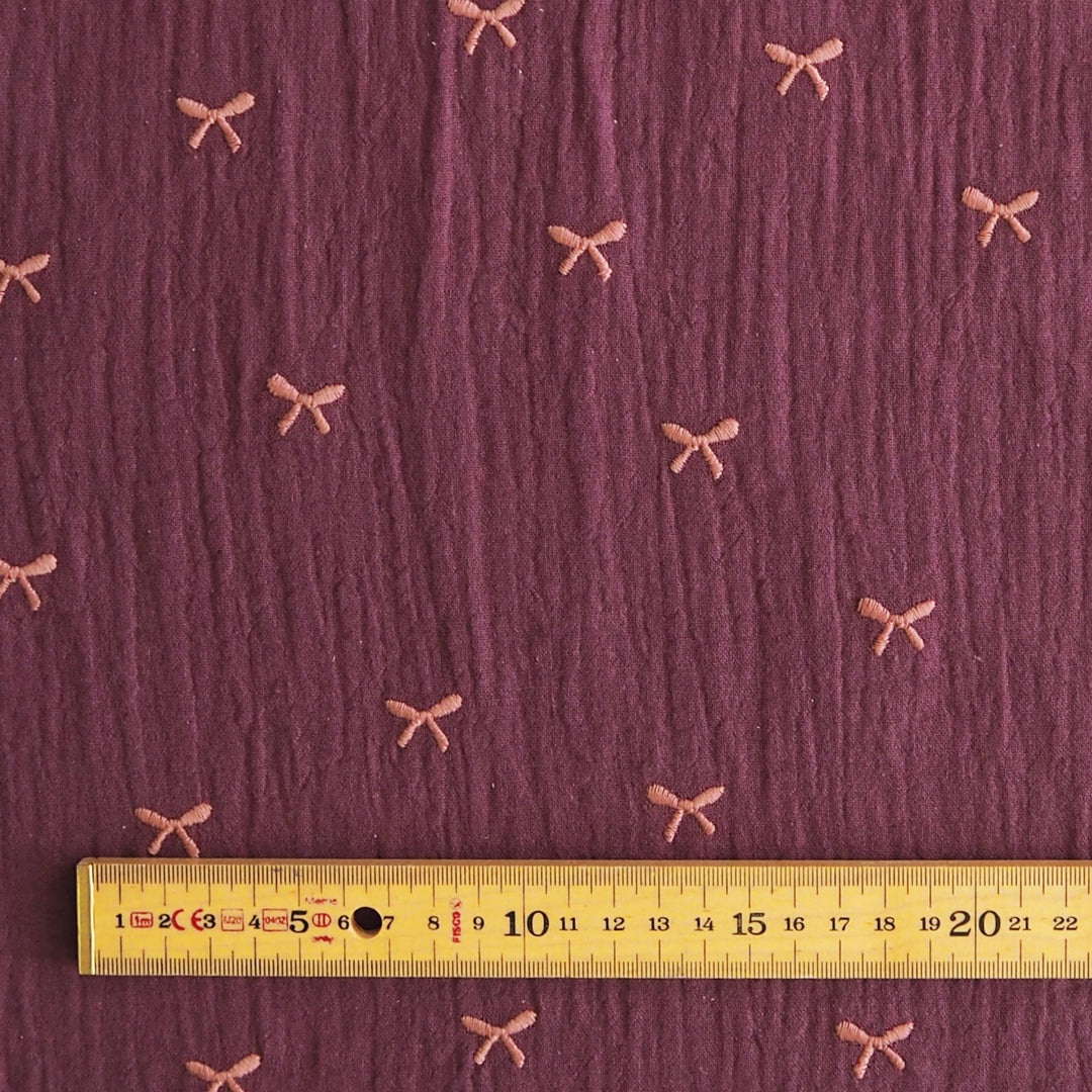 A close-up of textured mauve-purple fabric with small pink bow motifs repeated across it, with a yellow ruler along the bottom edge for scale.