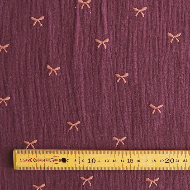 A close-up of textured mauve-purple fabric with small pink bow motifs repeated across it, with a yellow ruler along the bottom edge for scale.