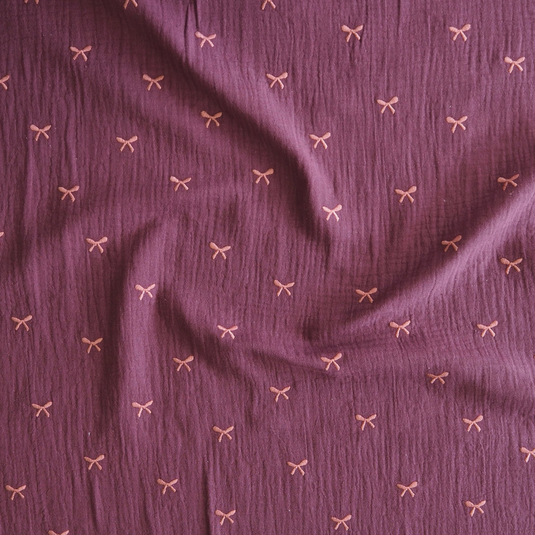 A close-up of textured mauve-purple fabric with a repeating small bow pattern in a light pinkish-beige tone.
