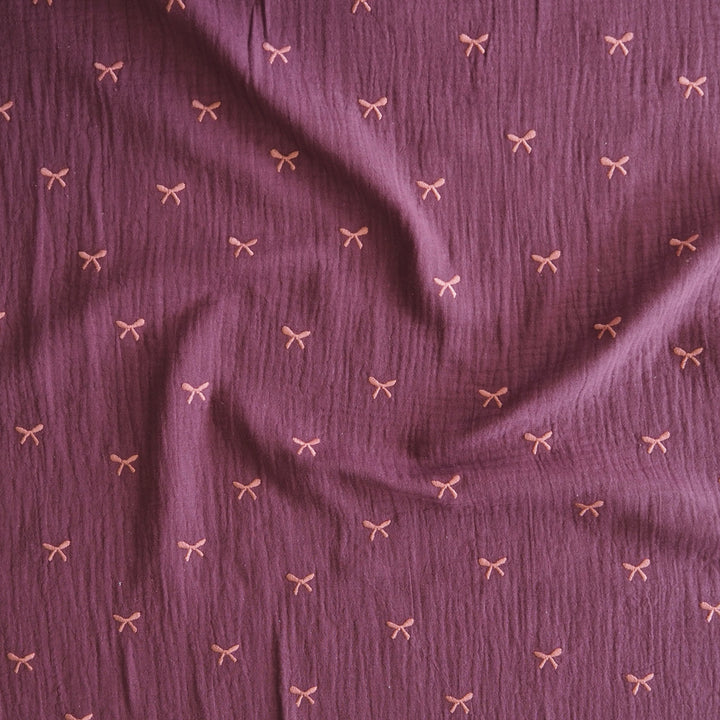A close-up of textured mauve-purple fabric with a repeating small bow pattern in a light pinkish-beige tone.