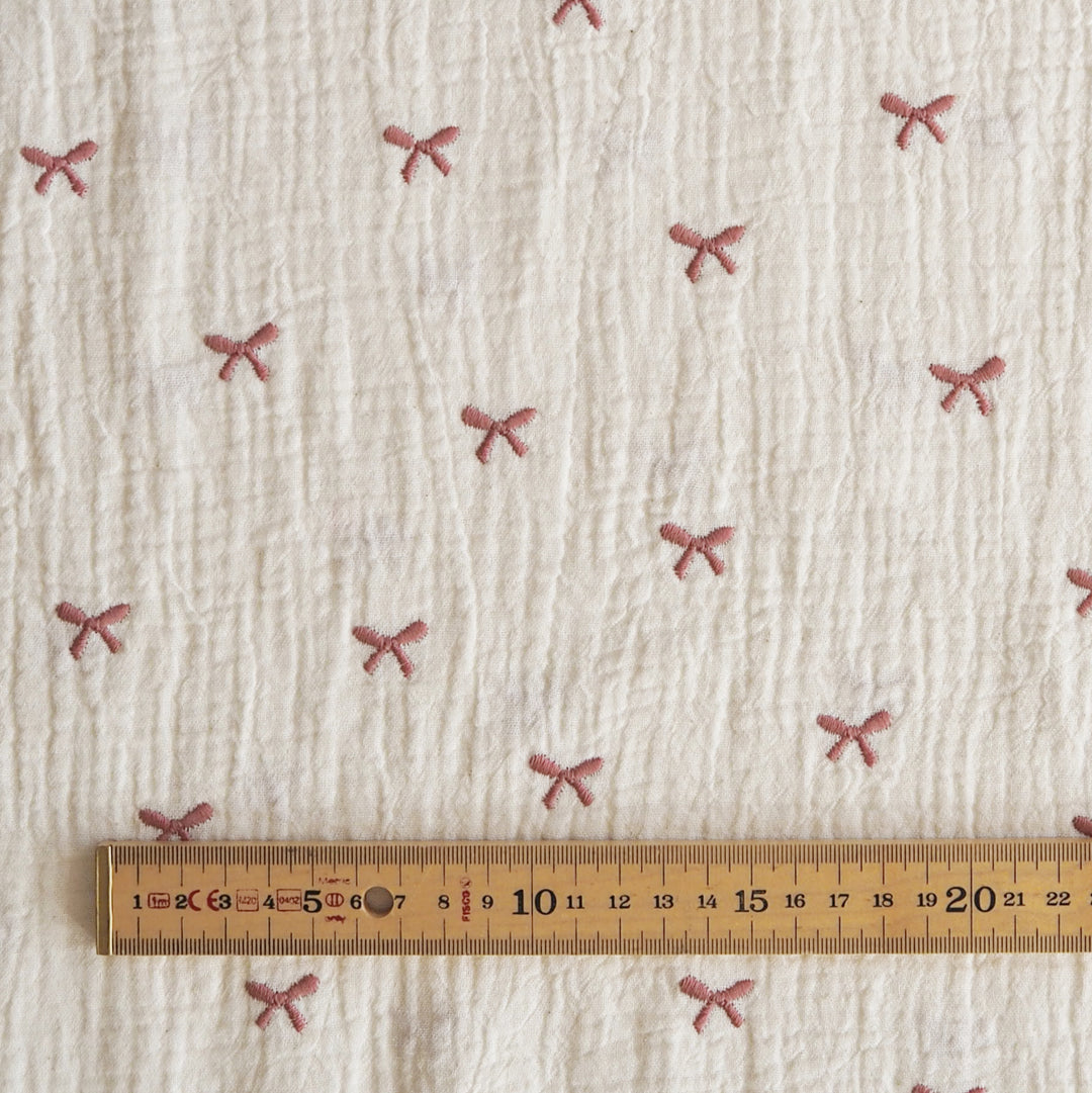 A close-up of textured off-white fabric with small pink bow motifs repeated across it, with a wooden ruler along the bottom edge for scale.
