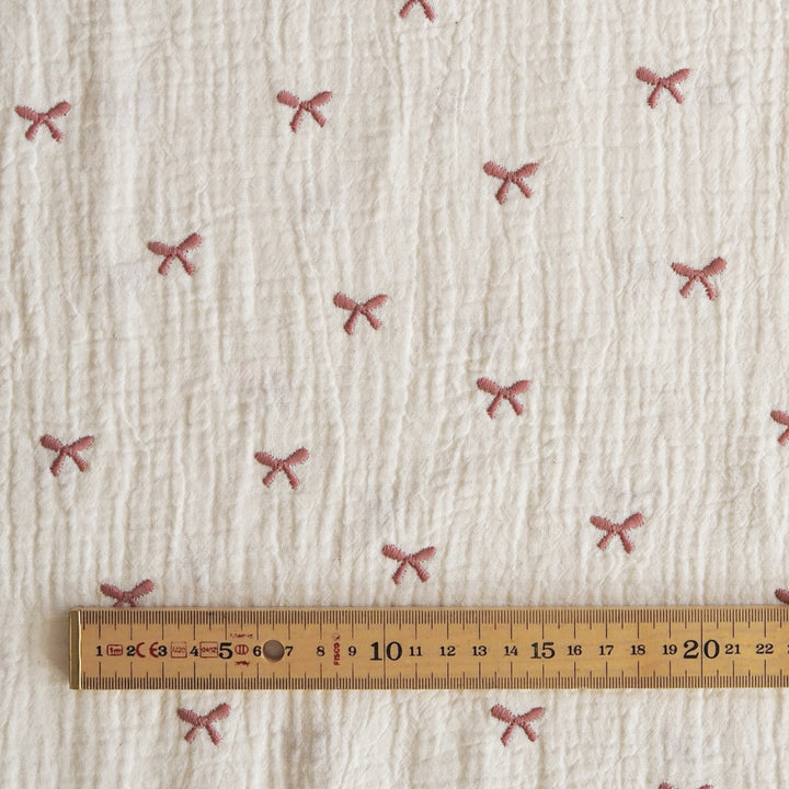 A close-up of textured off-white fabric with small pink bow motifs repeated across it, with a wooden ruler along the bottom edge for scale.