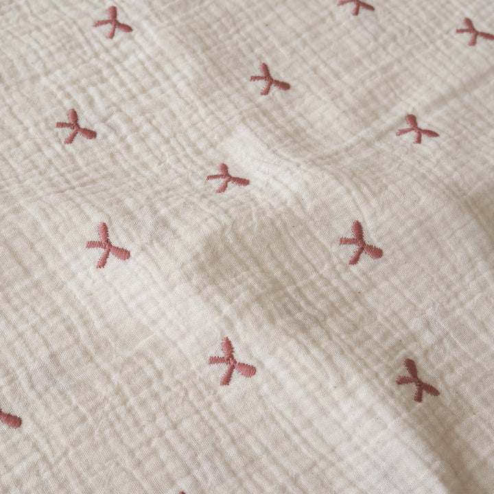 Close-up of a textured cream-colored muslin fabric with small, evenly spaced dusty pink embroidered cross motifs.