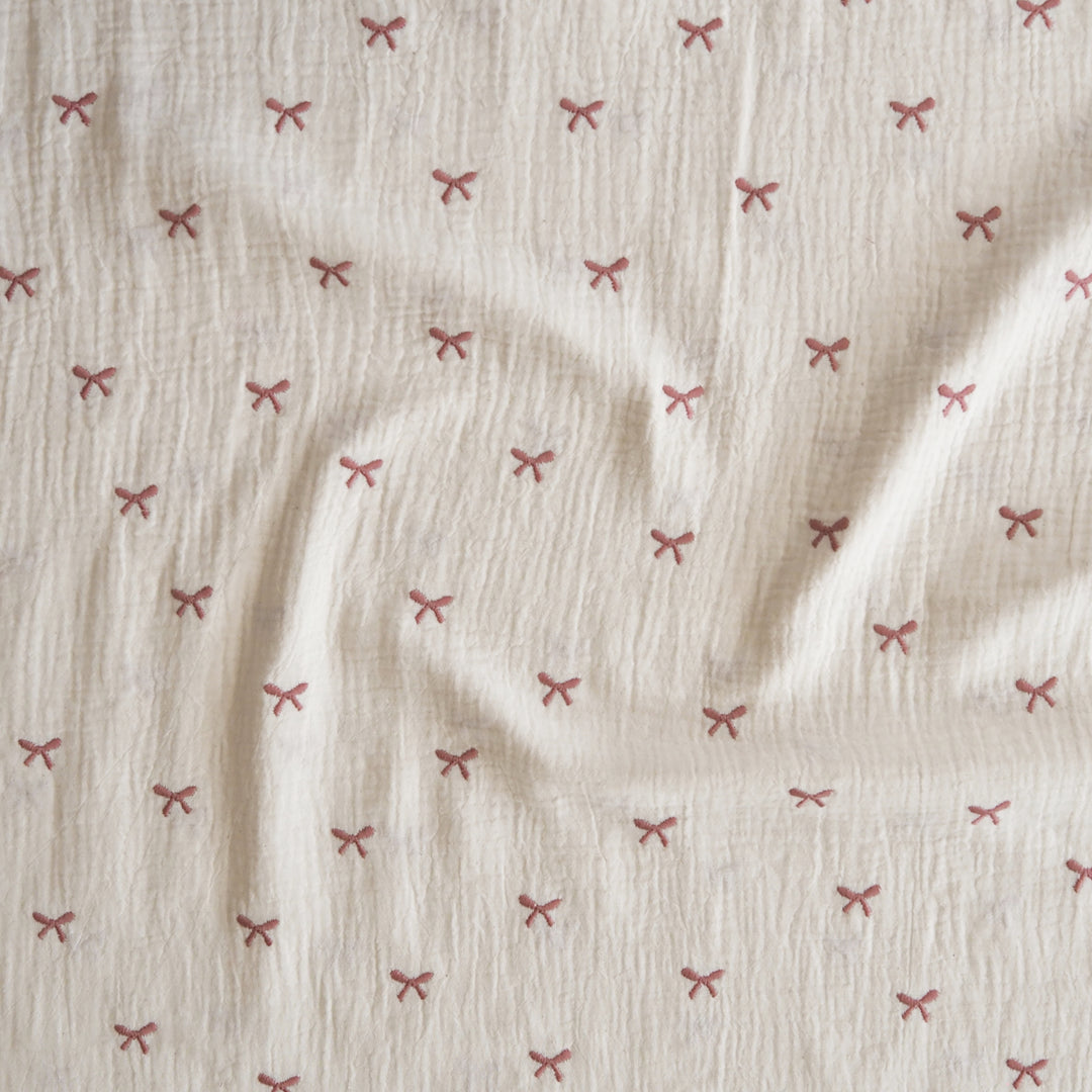 Off-white crinkled muslin fabric with a repeating pattern of small dusty pink embroidered bows, shown with soft folds.