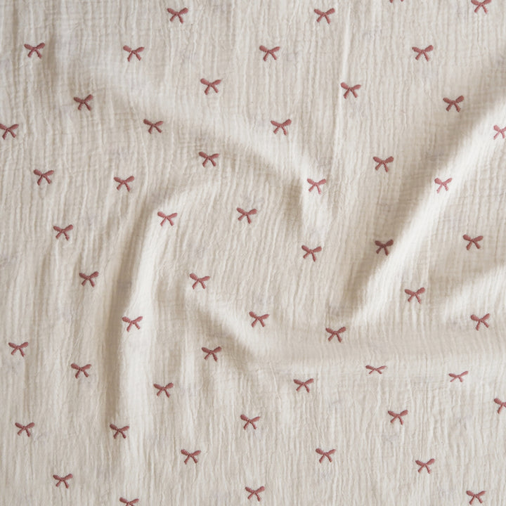Off-white crinkled muslin fabric with a repeating pattern of small dusty pink embroidered bows, shown with soft folds.