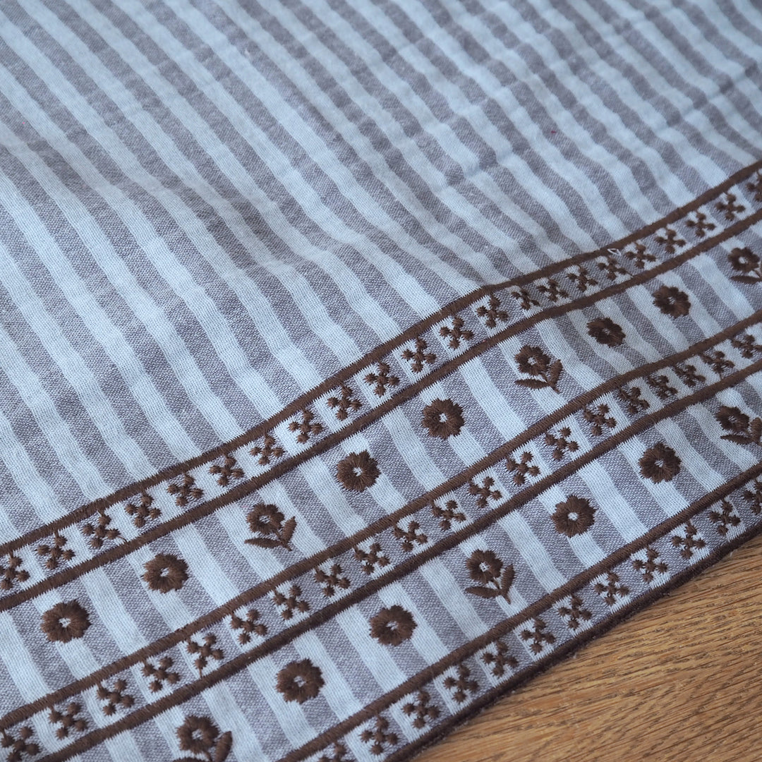 A close-up of a striped textile with a decorative border, laid on a wooden surface. The fabric has alternating light gray and medium gray vertical stripes, with a brown patterned band featuring floral motifs and geometric details along one edge.