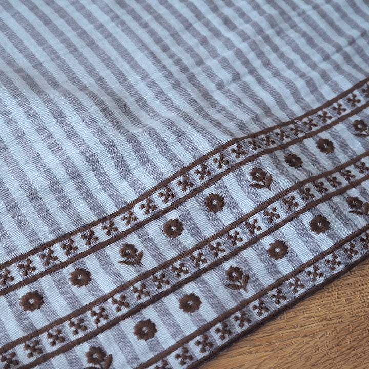 A close-up of a striped textile with a decorative border, laid on a wooden surface. The fabric has alternating light gray and medium gray vertical stripes, with a brown patterned band featuring floral motifs and geometric details along one edge.