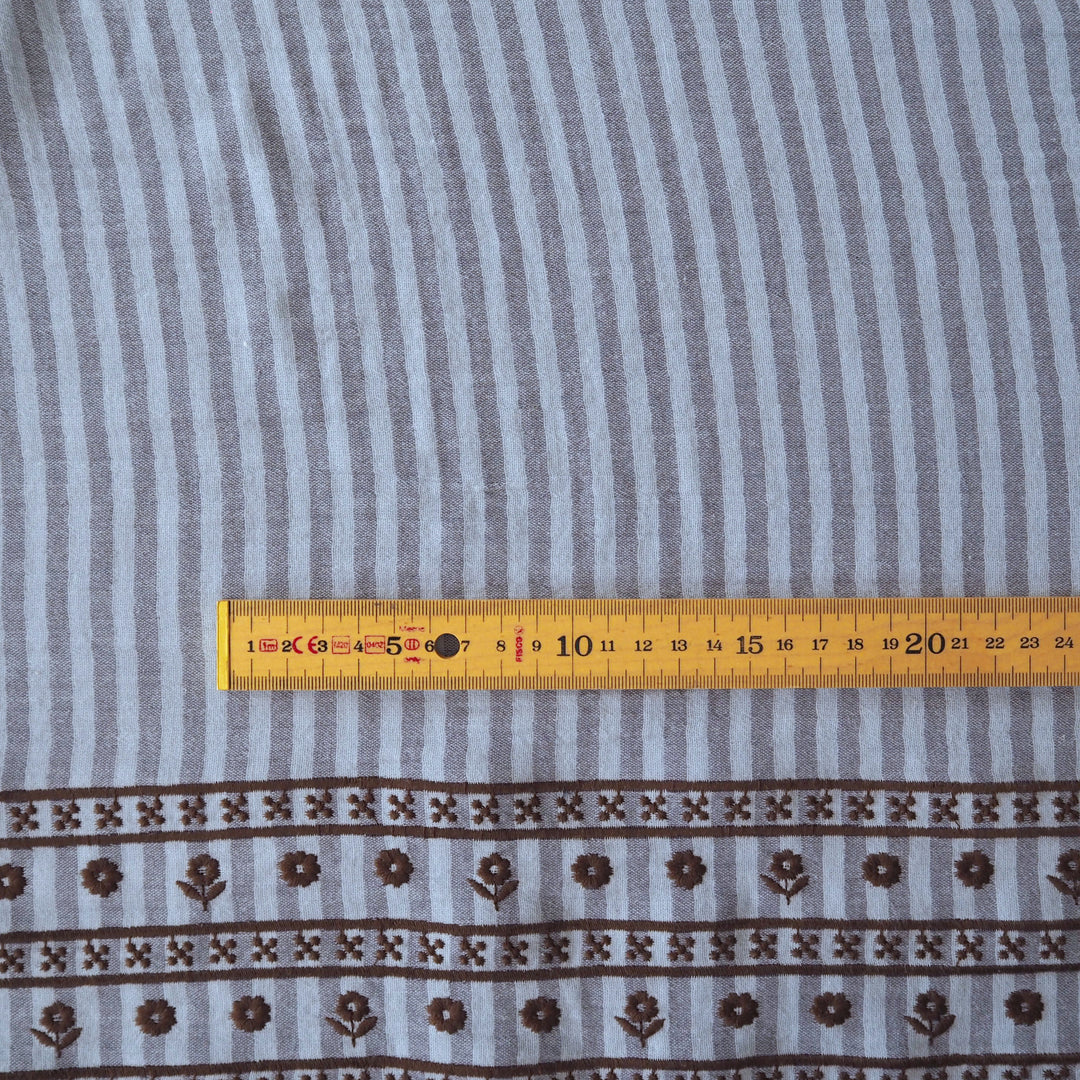 A close-up of a striped fabric with a brown floral border pattern, shown flat with a yellow measuring tape ruler across the middle for scale.