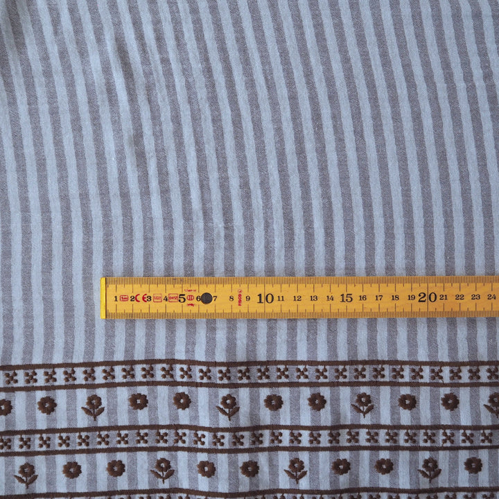 A close-up of a striped fabric with a brown floral border pattern, shown flat with a yellow measuring tape ruler across the middle for scale.