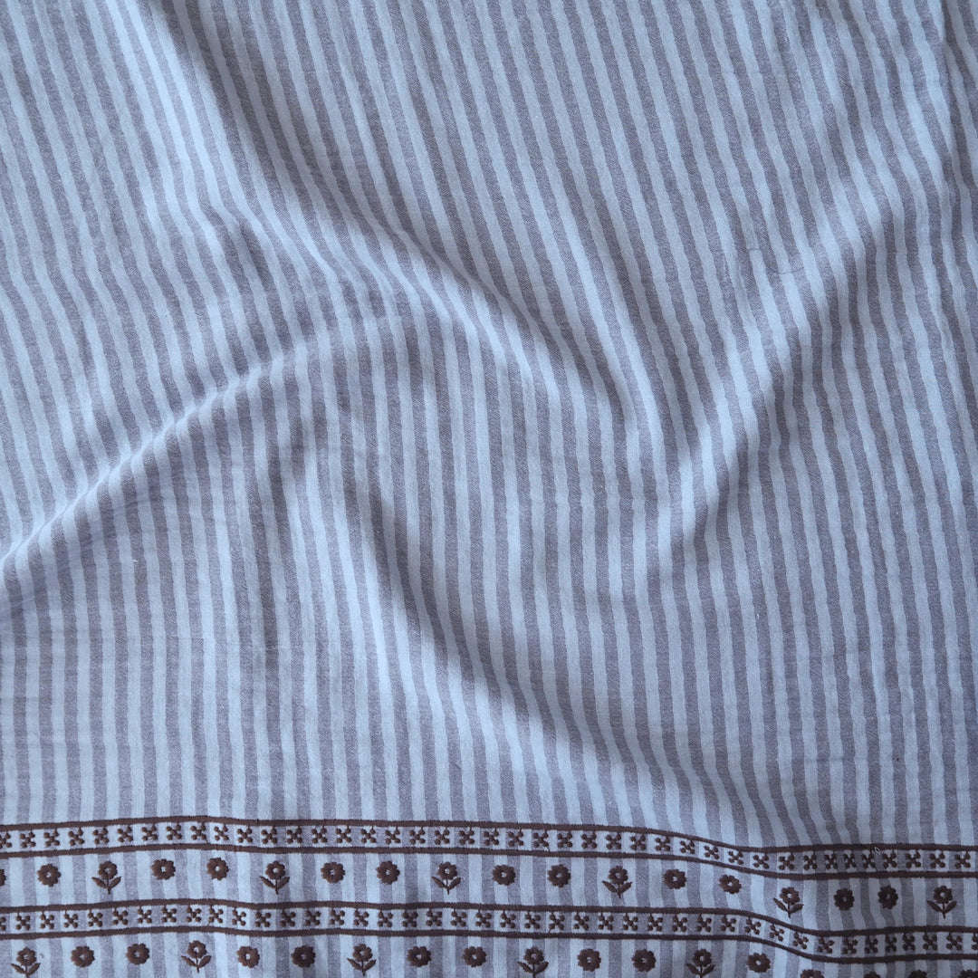 A close-up of a striped fabric featuring vertical light blue and white stripes with a decorative brown patterned border along the bottom edge.