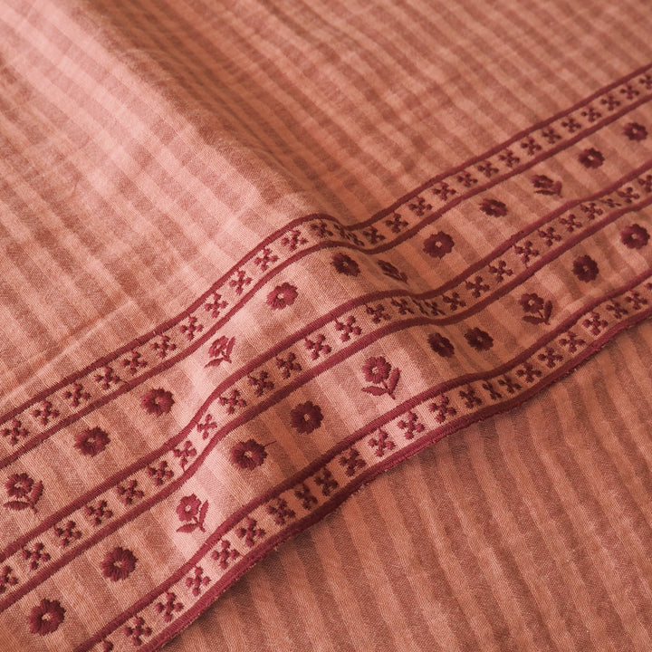 A close-up of a peach-colored woven fabric with a detailed border featuring floral and geometric patterns in a deep reddish-brown.