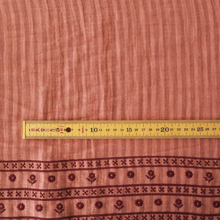 A close-up of a terracotta pink fabric with a vertical ribbed texture and a decorative border featuring dark red floral patterns, shown with a yellow measuring tape for scale.