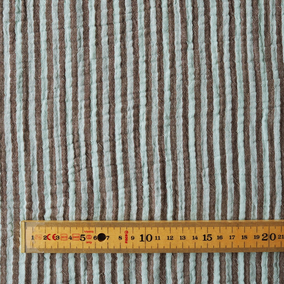 A close-up of a striped fabric with vertical brown and light gray-blue stripes, shown with a wooden measuring tape ruler along the bottom edge for scale.