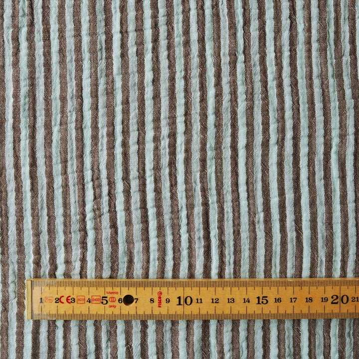 A close-up of a striped fabric with vertical brown and light gray-blue stripes, shown with a wooden measuring tape ruler along the bottom edge for scale.