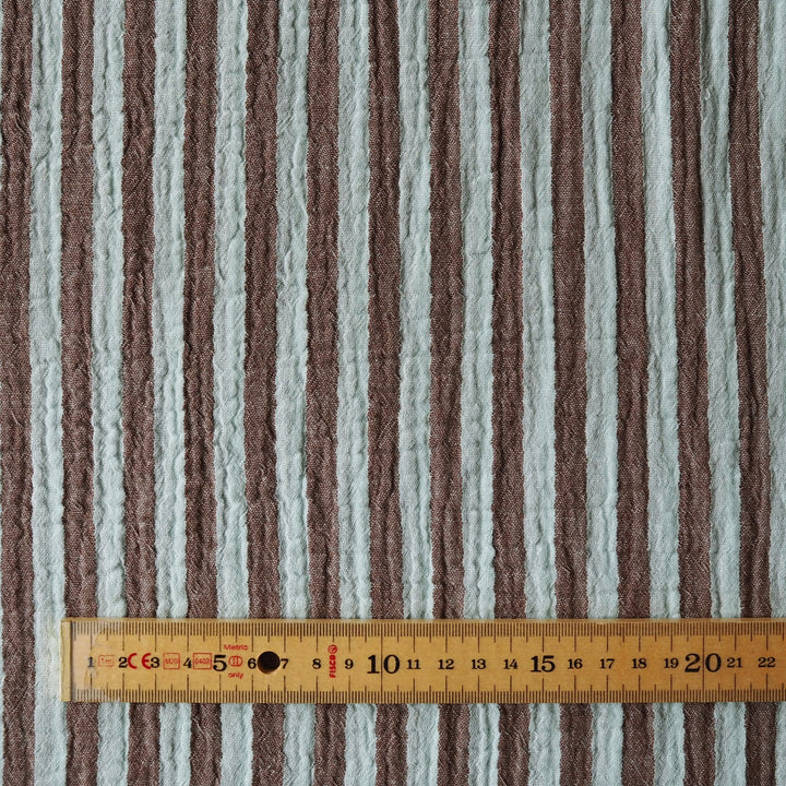A close-up of a striped fabric with alternating vertical bands of taupe-brown and light gray, shown with a wooden ruler at the bottom for scale.