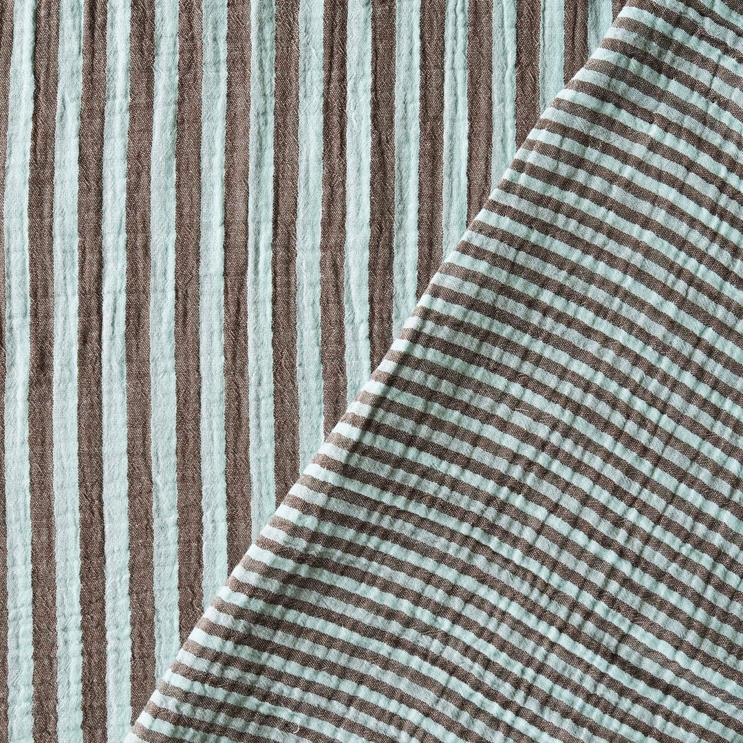 A close-up of a textured cotton muslin fabric with vertical brown and pale mint green stripes, folded diagonally to show its soft texture.