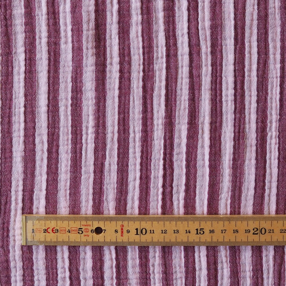 A close-up of a striped fabric with vertical wavy stripes in dusty mauve, light pink, and off-white, shown with a wooden ruler at the bottom for scale.