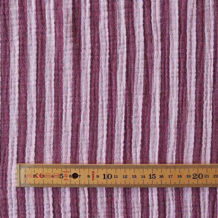 A close-up of a striped fabric with vertical wavy stripes in dusty mauve, light pink, and off-white, shown with a wooden ruler at the bottom for scale.