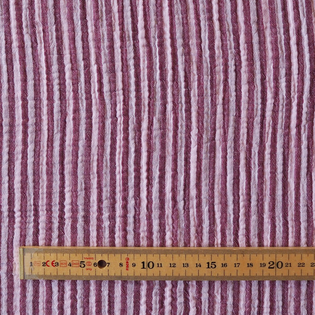 A close-up of a textured fabric with vertical stripes in dusty pink, mauve, and off-white, with a wooden ruler at the bottom for scale.