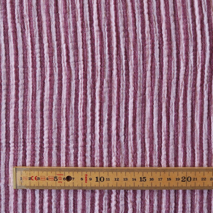 A close-up of a textured fabric with vertical stripes in dusty pink, mauve, and off-white, with a wooden ruler at the bottom for scale.