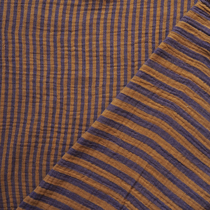 Close-up of a striped fabric with alternating vertical and diagonal bands in dark purple and warm mustard yellow, showing a textured, gauze-like weave.