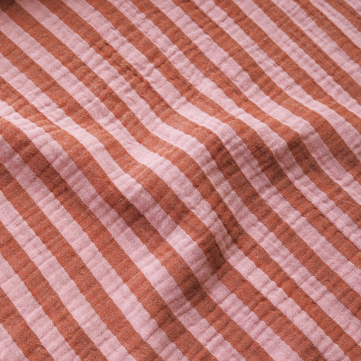 A close-up of a textured, double-gauze cotton fabric with diagonal stripes in terracotta orange and dusty pink.