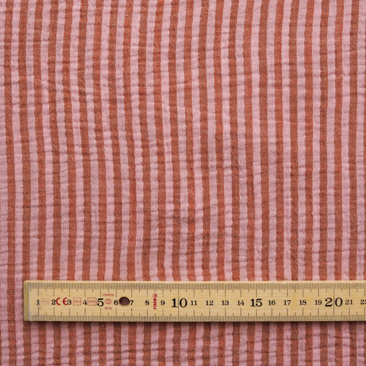 A close-up of a pink and rust-red vertically striped fabric with a wooden ruler along the bottom edge for scale.