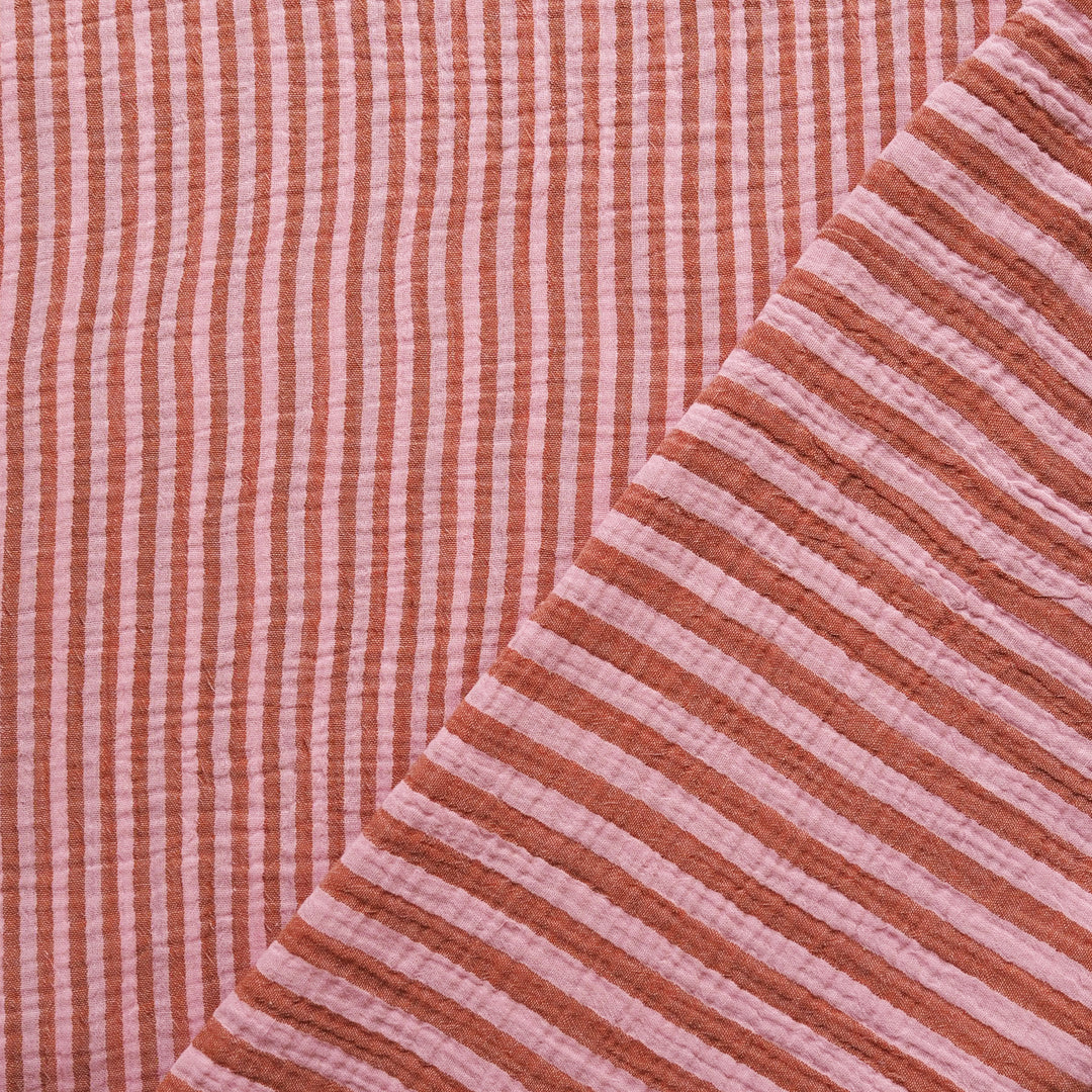 Close-up of a textured muslin fabric with vertical pink and terracotta stripes, folded diagonally to show its soft, crinkled texture.