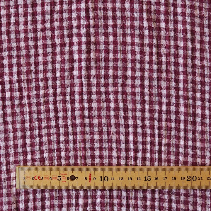 A close-up of a red and white gingham patterned fabric, with a wooden ruler placed along the bottom edge to show scale.