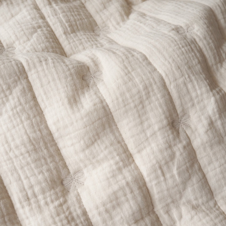 Close-up of a textured cream-colored muslin blanket with stitched bow details, showing its soft, crinkled fabric and quilted folds.