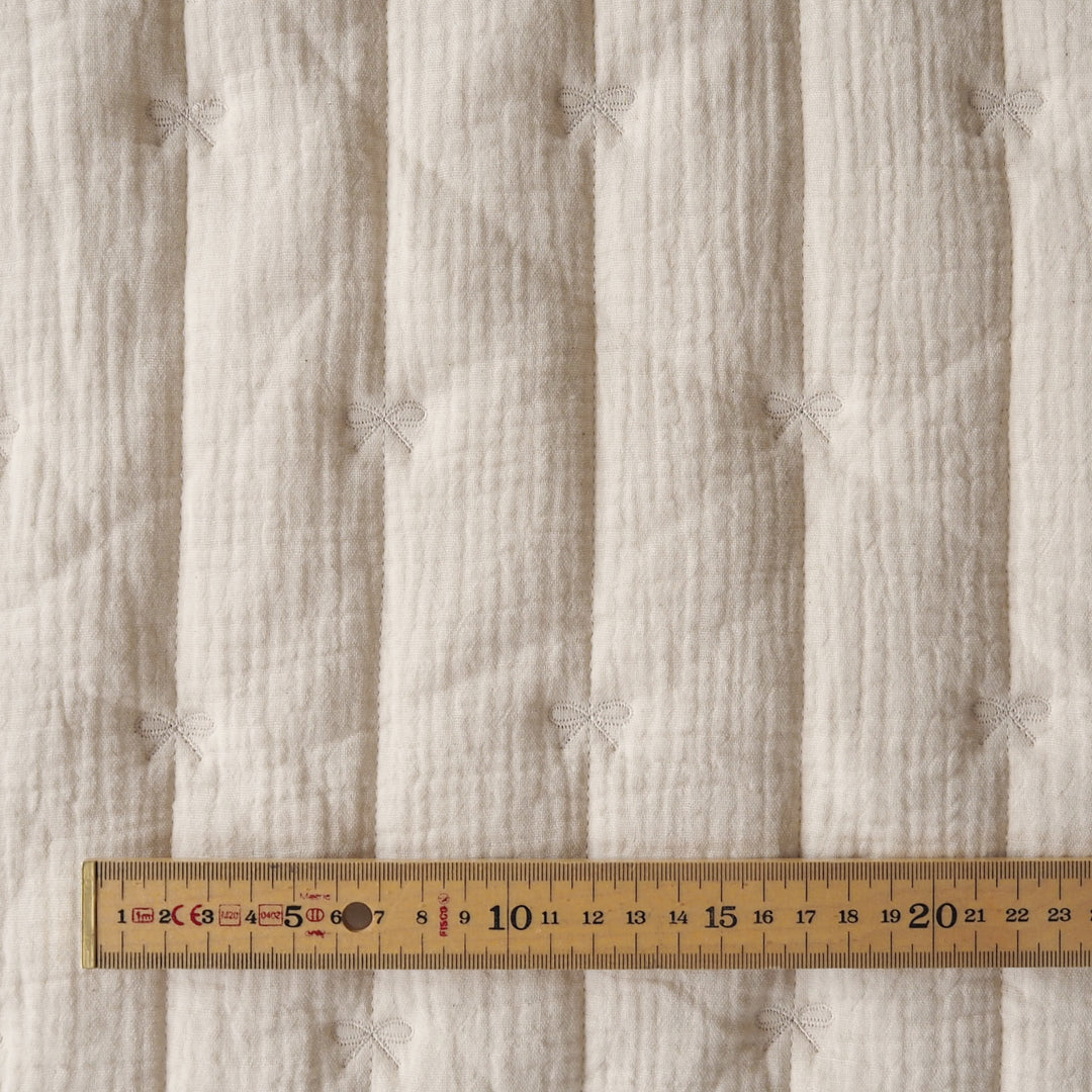 Close-up of a textured off-white quilted fabric with vertical channels and small bow details, shown with a wooden ruler along the bottom to indicate scale.