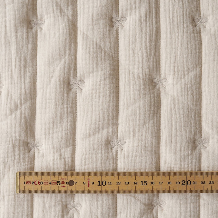 Close-up of a textured off-white quilted fabric with vertical channels and small bow details, shown with a wooden ruler along the bottom to indicate scale.
