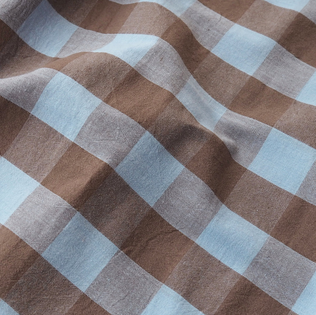 A close-up of a soft woven fabric with a brown and light blue gingham plaid pattern, shown with gentle folds.