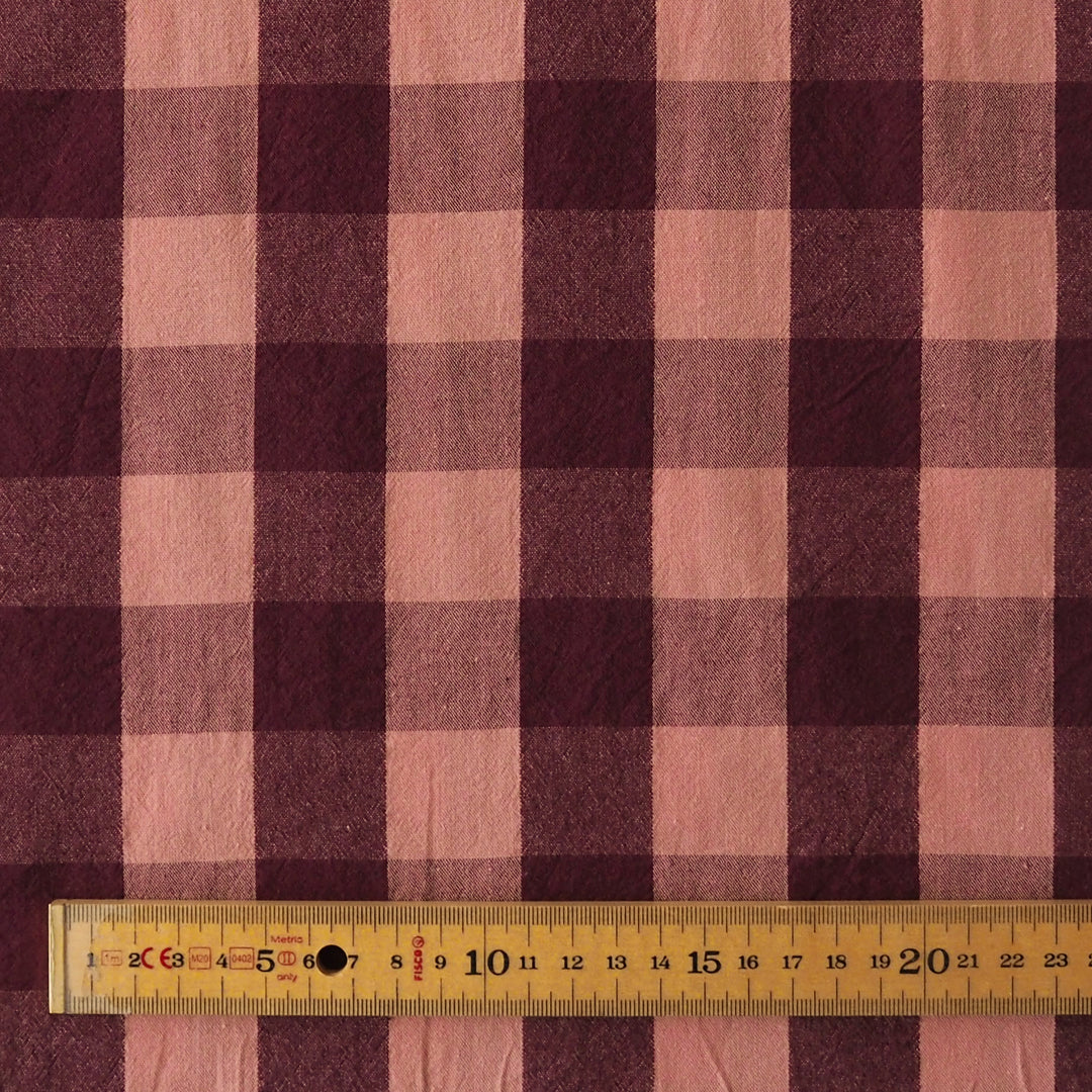 A close-up of a plaid fabric pattern in dusty pink and dark burgundy, with a wooden ruler at the bottom showing the scale of the check pattern.