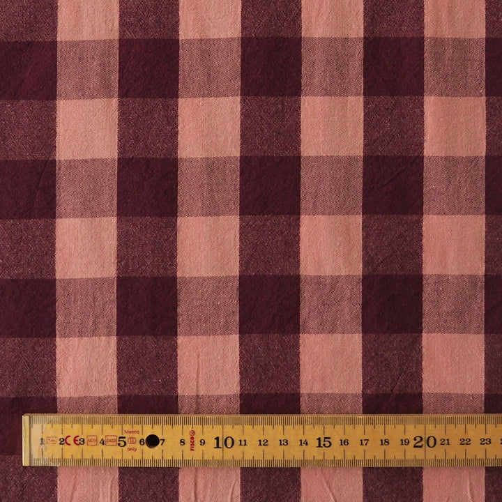 A close-up of a plaid fabric pattern in dusty pink and dark burgundy, with a wooden ruler at the bottom showing the scale of the check pattern.