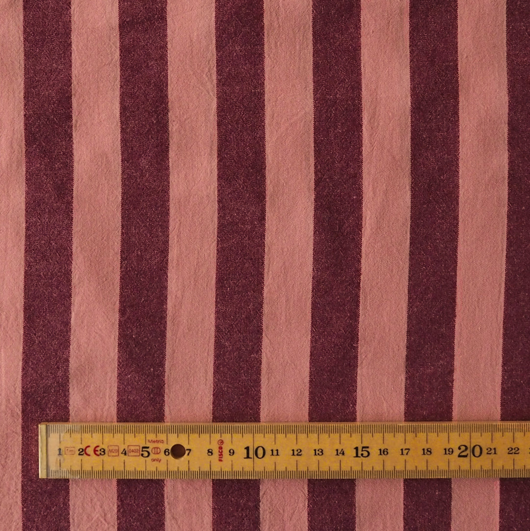 A close-up of striped fabric with alternating dusty pink and dark burgundy vertical stripes, with a yellow metal ruler laid horizontally near the bottom to show scale.