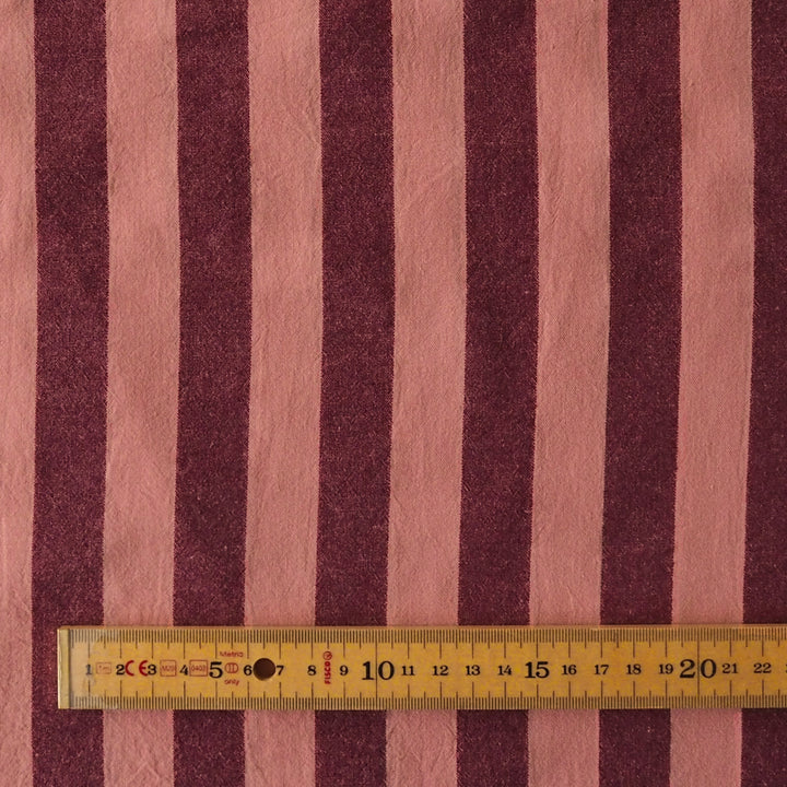 A close-up of striped fabric with alternating dusty pink and dark burgundy vertical stripes, with a yellow metal ruler laid horizontally near the bottom to show scale.