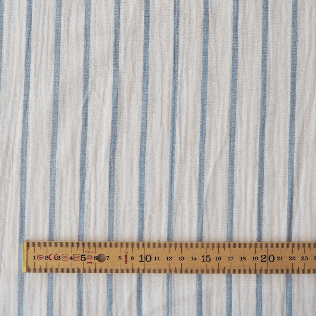 A close-up of a white fabric with vertical light blue stripes, shown with a wooden ruler at the bottom to indicate scale.
