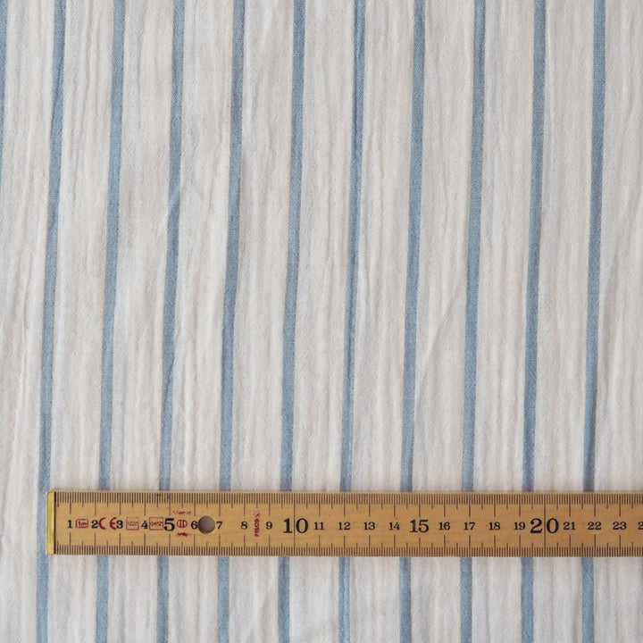 A close-up of a white fabric with vertical light blue stripes, shown with a wooden ruler at the bottom to indicate scale.