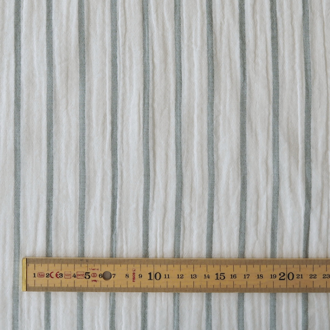A close-up of a white fabric with vertical gray stripes, shown with a wooden ruler at the bottom to indicate scale.