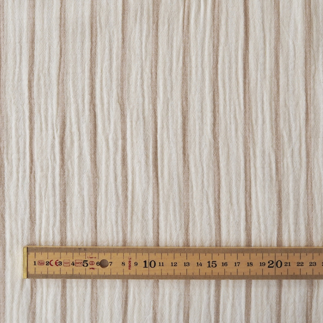 A close-up of a textured fabric or wallpaper with vertical beige and off-white stripes, shown with a wooden ruler at the bottom for scale.