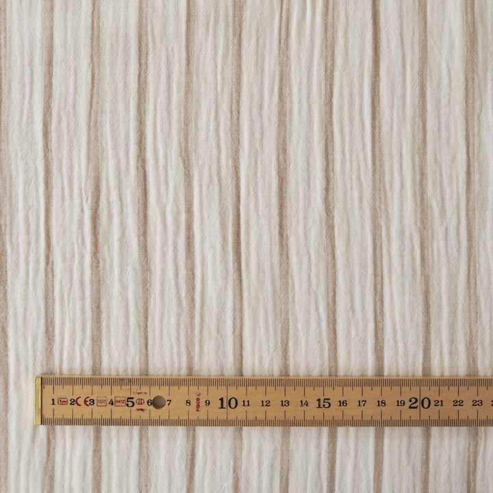 A close-up of a textured fabric or wallpaper with vertical beige and off-white stripes, shown with a wooden ruler at the bottom for scale.