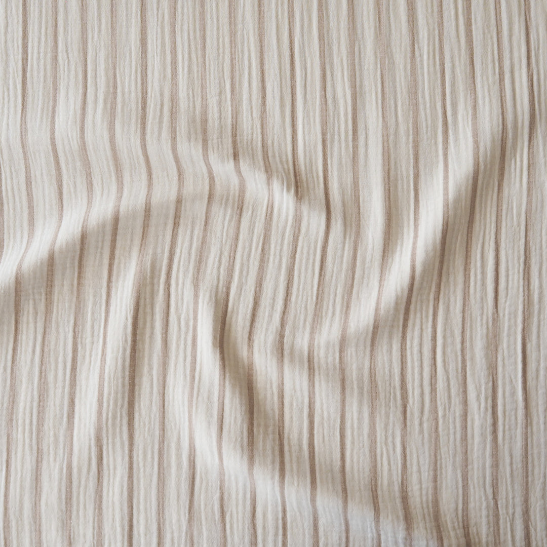 A close-up of a textured fabric with vertical beige and cream stripes, showing a soft crinkled or gauze-like surface with gentle folds.