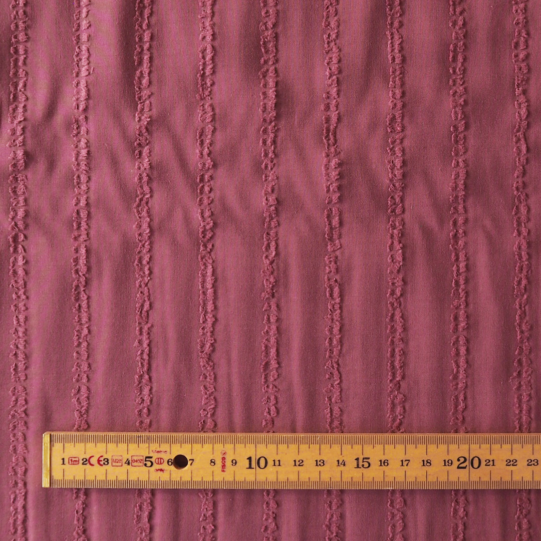 A close-up of a dusty rose pink textured fabric with vertical raised stripes, shown with a wooden ruler at the bottom for scale.