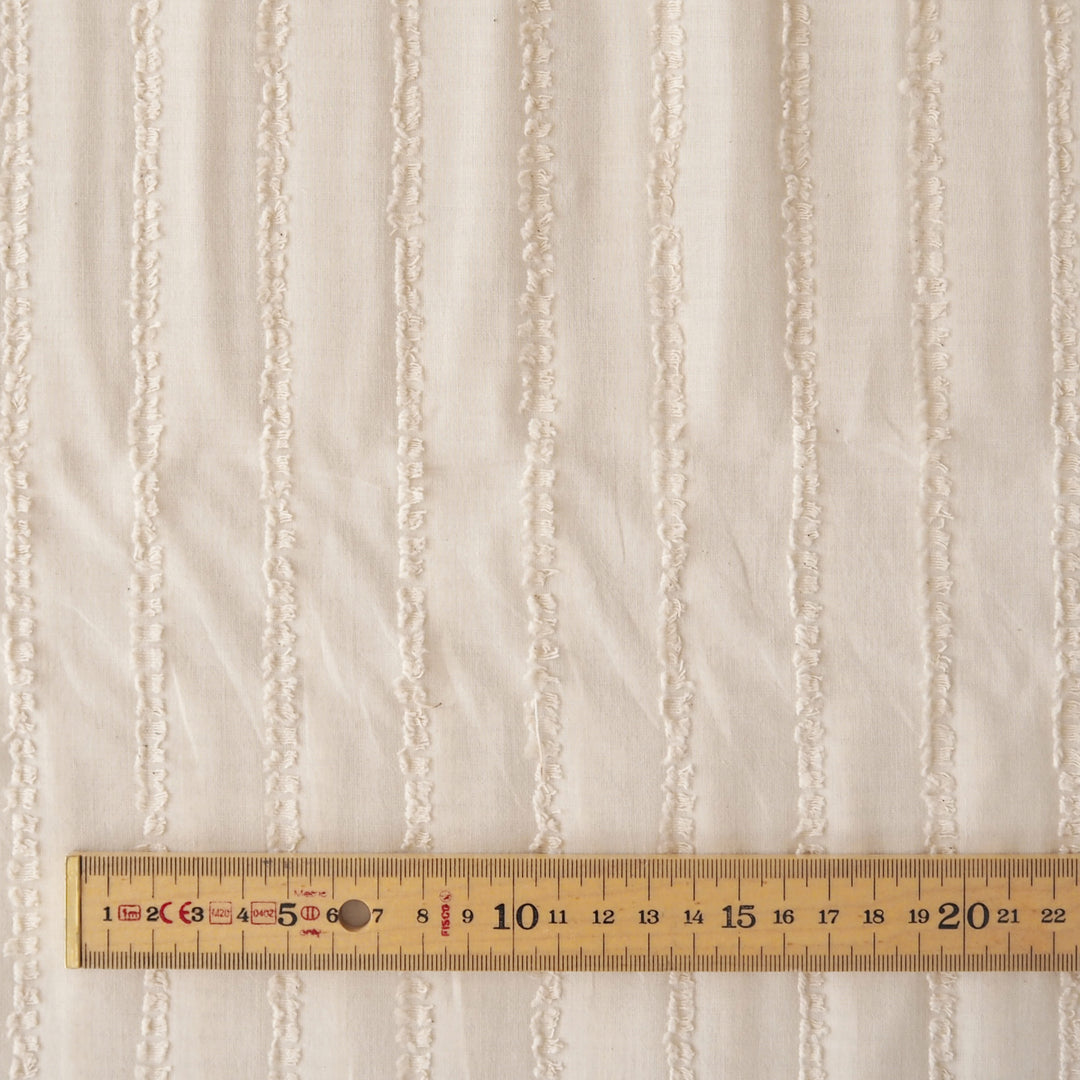 Close-up of a textured cream fabric with vertical ribbed stripes, shown with a wooden ruler at the bottom to indicate scale.