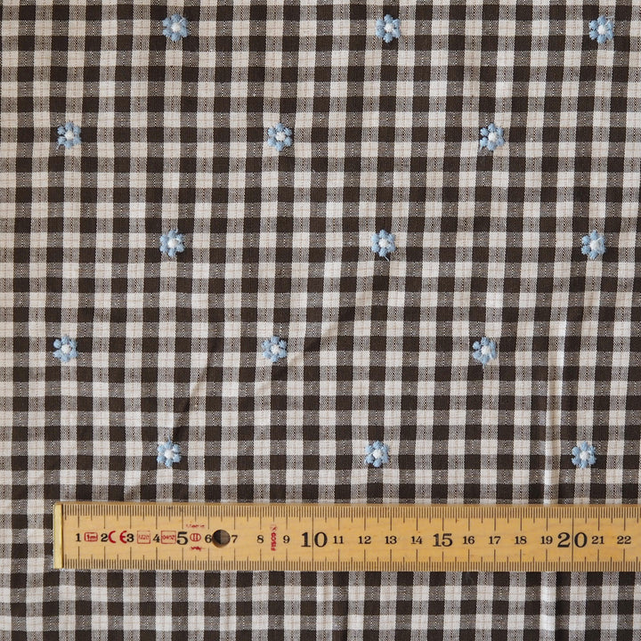 Brown and beige gingham fabric with small light blue embroidered flowers, shown flat with a wooden ruler at the bottom for scale.