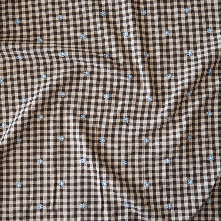 Brown and white gingham fabric with small light blue embroidered floral motifs, shown in a draped, wrinkled state.