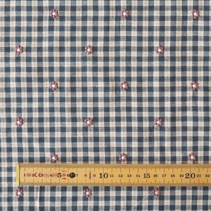 A close-up of a blue and beige gingham fabric with small pink and white embroidered floral motifs, shown with a yellow measuring tape along the bottom edge for scale.